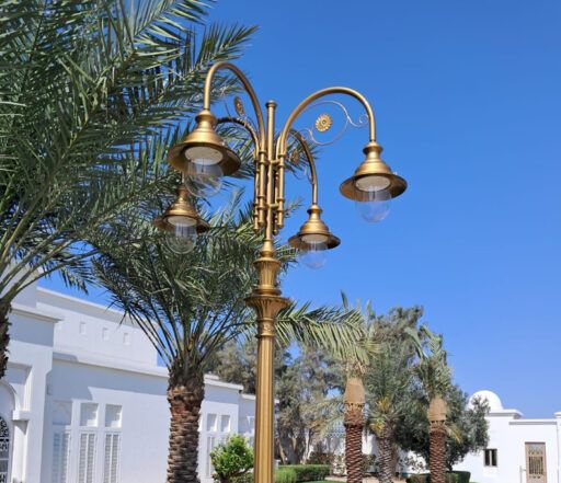 Farola ricamente decorada con pantalla dorada y cuatro luminarias 04/Aries/A, sobre un fondo de palmeras y edificios blancos en un día soleado.